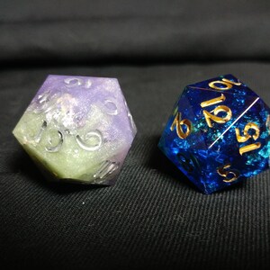May include: Two iridescent dice, one purple and green with silver numbers, the other blue with gold numbers.