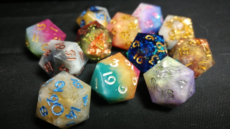 May include: A collection of ten multi-colored, iridescent, and glittery dice with white numbers. The dice are made of resin and have a variety of colors, including blue, green, pink, purple, and gold.
