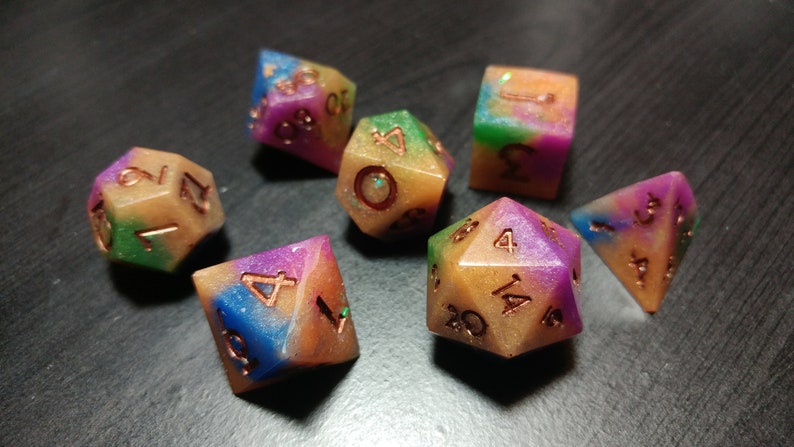 Puede incluir: Un juego de siete dados poli&eacute;dricos con un aspecto colorido y veteado. Los dados presentan una mezcla de colores que incluyen azul, morado, verde y dorado. Los n&uacute;meros son de color cobre. Ideales para juegos de mesa.