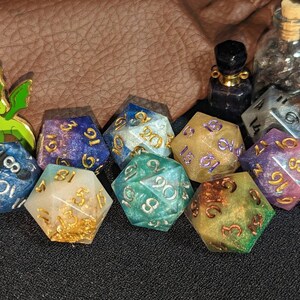 Puede incluir: Una colecci&oacute;n de dados poli&eacute;dricos de m&uacute;ltiples caras en varios colores, incluyendo azul, morado, verde y dorado, con n&uacute;meros met&aacute;licos. El conjunto incluye una peque&ntilde;a figura de drag&oacute;n verde, dos viales de vidrio y un colgante de metal.