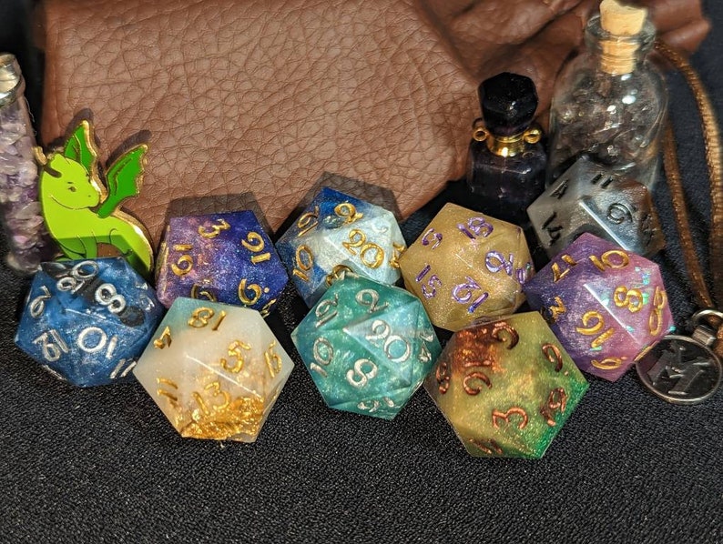Puede incluir: Una colecci&oacute;n de dados poli&eacute;dricos de m&uacute;ltiples caras en varios colores, incluyendo azul, morado, verde y dorado, con n&uacute;meros met&aacute;licos. El conjunto incluye una peque&ntilde;a figura de drag&oacute;n verde, dos viales de vidrio y un colgante de metal.