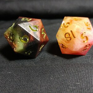 May include: Two multi-colored dice with gold numbers. The dice are made of resin and have a speckled appearance. One die is black, green, and red, and the other is pink and white.