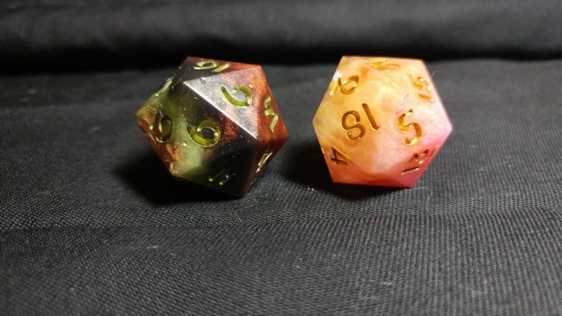 May include: Two multi-colored dice with gold numbers. The dice are made of resin and have a speckled appearance. One die is black, green, and red, and the other is pink and white.