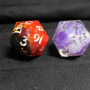 Pu&ograve; includere: Due dadi, uno rosso e nero con macchie dorate, l'altro viola e bianco con glitter. Entrambi i dadi hanno numeri bianchi.
