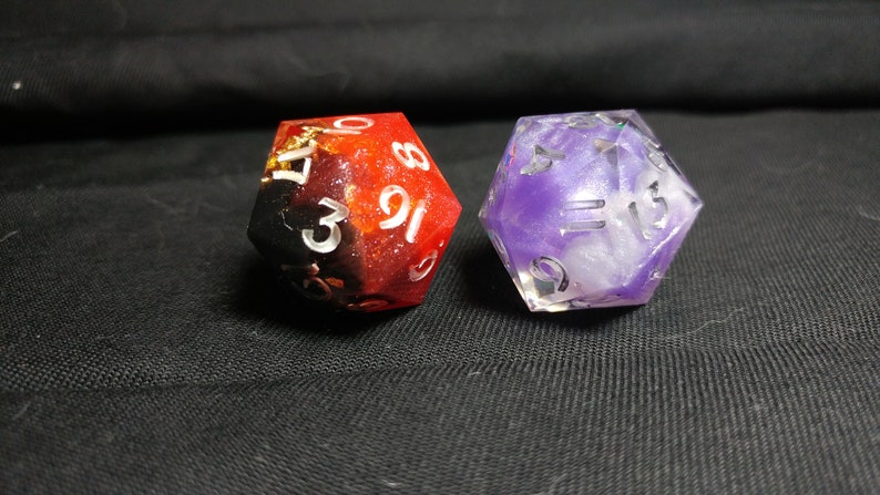 Pu&ograve; includere: Due dadi, uno rosso e nero con macchie dorate, l'altro viola e bianco con glitter. Entrambi i dadi hanno numeri bianchi.