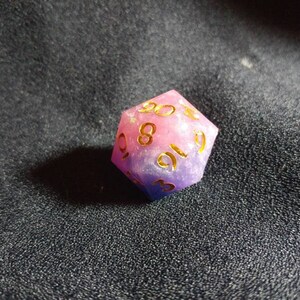 Puede incluir: Un dado poli&eacute;drico de veinte caras que presenta una mezcla de resina rosa y morada con inclusiones brillantes. Los n&uacute;meros est&aacute;n grabados en la superficie y rellenos con un pigmento dorado met&aacute;lico, lo que proporciona un contraste claro.