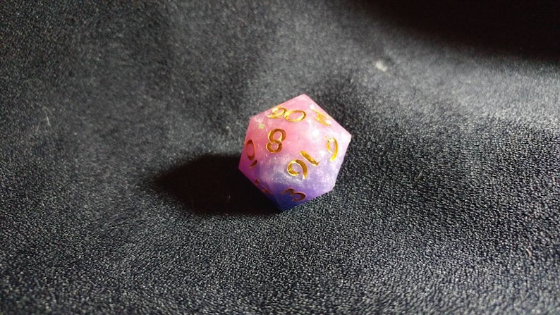 Puede incluir: Un dado poli&eacute;drico de veinte caras que presenta una mezcla de resina rosa y morada con inclusiones brillantes. Los n&uacute;meros est&aacute;n grabados en la superficie y rellenos con un pigmento dorado met&aacute;lico, lo que proporciona un contraste claro.