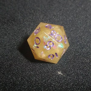 Puede incluir: Un dado de veinte caras hecho de resina amarilla transl&uacute;cida con escamas de purpurina iridiscente incrustadas. El dado presenta n&uacute;meros grabados pintados en color morado oscuro, incluyendo los n&uacute;meros 20, 16, 10, 8 y 2 en la superficie.
