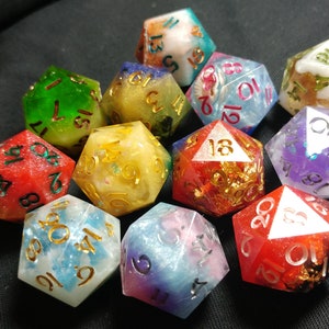 Pu&ograve; includere: Un set di dadi a 12 facce con vari colori e accenti glitter. I dadi sono realizzati in resina e hanno numeri dorati. I dadi sono disposti in una pila su una superficie nera.
