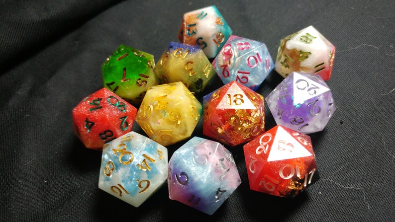 Pu&ograve; includere: Un set di dadi a 12 facce con vari colori e accenti glitter. I dadi sono realizzati in resina e hanno numeri dorati. I dadi sono disposti in una pila su una superficie nera.