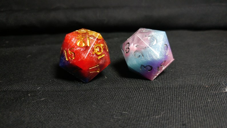 Pu&ograve; includere: Due dadi multi-colorati e iridescenti in resina con numeri dorati. Il dado a sinistra &egrave; rosso e blu, mentre quello a destra &egrave; blu, rosa e viola.