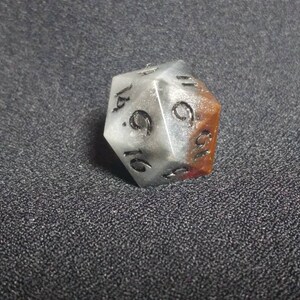 Puede incluir: Un dado de veinte caras con un cuerpo transl&uacute;cido de color blanco y gris jaspeado, con una secci&oacute;n de color marr&oacute;n rojizo en un lado. Los n&uacute;meros est&aacute;n grabados en negro, con el n&uacute;mero seis claramente visible en la cara triangular frontal.