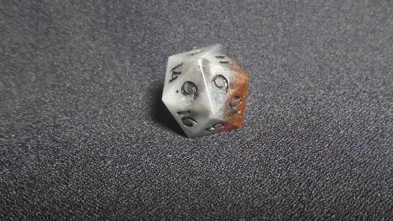 Puede incluir: Un dado de veinte caras con un cuerpo transl&uacute;cido de color blanco y gris jaspeado, con una secci&oacute;n de color marr&oacute;n rojizo en un lado. Los n&uacute;meros est&aacute;n grabados en negro, con el n&uacute;mero seis claramente visible en la cara triangular frontal.