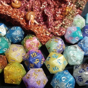 Puede incluir: Un conjunto de dados multicolores de m&uacute;ltiples caras, con n&uacute;meros dorados y plateados, colocados frente a una bolsa de punto. Los dados son de varios colores, incluyendo azul, verde, morado y amarillo. La bolsa es marr&oacute;n y roja.