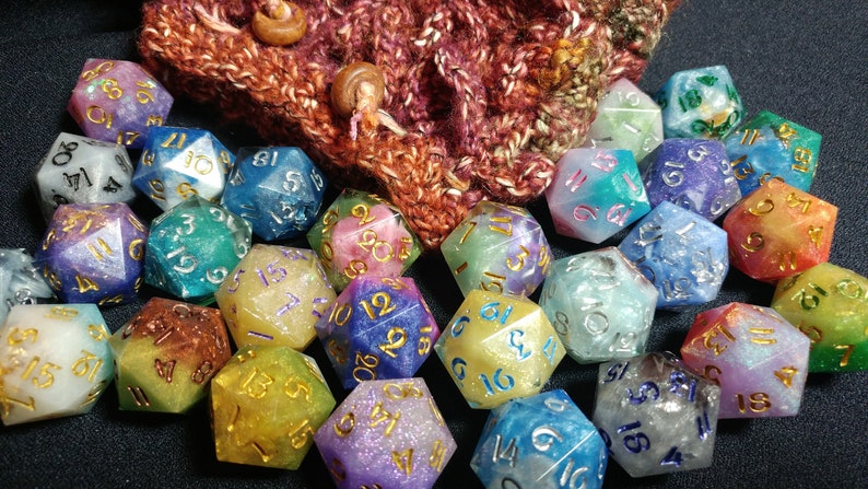 Puede incluir: Un conjunto de dados multicolores de m&uacute;ltiples caras, con n&uacute;meros dorados y plateados, colocados frente a una bolsa de punto. Los dados son de varios colores, incluyendo azul, verde, morado y amarillo. La bolsa es marr&oacute;n y roja.