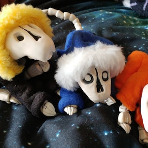 May include: Three plush skeleton figures with white bone structures and fabric clothing. One has yellow fur, black clothing, and a white bone tail. Another has a blue hat with white trim and orange clothing. The third has orange clothing.