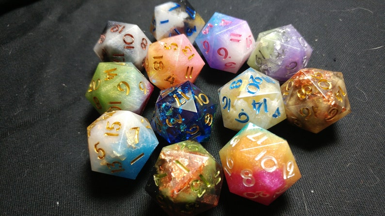 May include: A set of 12 iridescent dice with gold numbers. The dice are made of resin and have a variety of colors, including blue, green, pink, and purple. Each die has a different color and pattern.