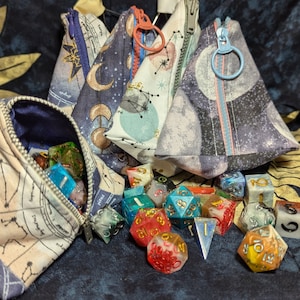 May include: Three fabric pouches with zipper closures and celestial print designs. The pouches are filled with colorful dice. The pouches are on a blue and gold patterned fabric.