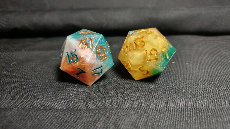 Pu&ograve; includere: Due dadi multicolori con numeri dorati. I dadi sono in resina e hanno una finitura scintillante. Un dado &egrave; turchese, bianco e rame, e l'altro &egrave; giallo, bianco e verde.