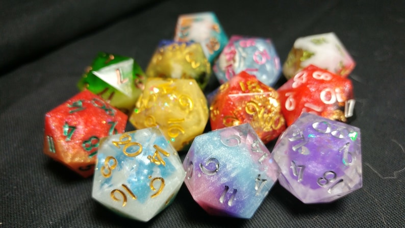 Pu&ograve; includere: Un set di dadi a dieci facce con glitter iridescente incorporato nella resina. I dadi sono di vari colori, tra cui rosso, verde, blu, viola e giallo. Ogni dado ha numeri bianchi sulle facce.