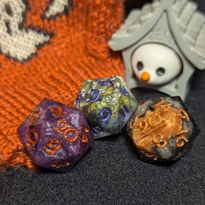 Puede incluir: Tres dados iridiscentes con diferentes colores y purpurina. Los dados son de color morado oscuro, verde y dorado, y negro y dorado. Los dados están sobre una superficie gris oscuro.