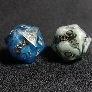 Puede incluir: Dos dados de veinte caras hechos de resina. Un dado es de color azul intenso con remolinos de tonos azules m&aacute;s claros, y el otro es de color gris p&aacute;lido con patrones de m&aacute;rmol blanco. Ambos dados tienen n&uacute;meros plateados grabados.