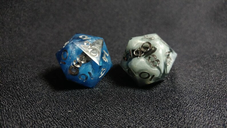 Puede incluir: Dos dados de veinte caras hechos de resina. Un dado es de color azul intenso con remolinos de tonos azules m&aacute;s claros, y el otro es de color gris p&aacute;lido con patrones de m&aacute;rmol blanco. Ambos dados tienen n&uacute;meros plateados grabados.