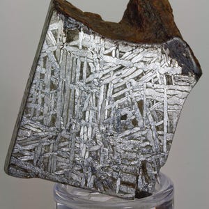 May include: A polished, angular meteorite fragment with a Widmanstätten pattern, showcasing a complex, interwoven crystalline structure. The metallic silver interior contrasts with the dark, oxidized exterior. The specimen is displayed on a clear, circular stand.