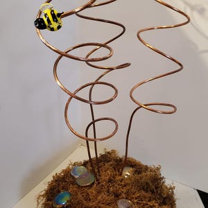 May include: A copper wire sculpture with three spiraling branches, each topped with a glass insect. The insects are a red ladybug, a blue butterfly, and a yellow and black bee. The sculpture is set in a brown moss base with iridescent glass stones.