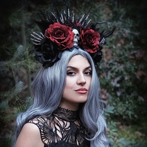May include: A woman with long, silver hair wears a black lace top and a dramatic crown made of black feathers, red roses, and a skull. The crown is adorned with black and silver skeletal hands.