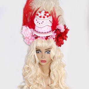 May include: A mannequin head with long, wavy blonde hair and a decorative headpiece. The headpiece features a pink cake, red feathers, and pink and red flowers. The mannequin has blue eyes and red lipstick.