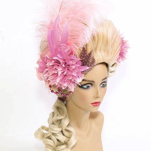 May include: A blonde wig with a large pink flower and feather embellishment. The wig is styled in a high, bouffant style with a long, curled ponytail.
