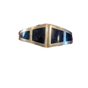 Könnte beinhalten: Ein silbernes Armband mit vier eingelegten Segmenten aus dunkelblauem Stein mit sichtbarer natürlicher Maserung. Das Metallband hat eine glatte, matte Oberfläche und ein geometrisches, sich verjüngendes Design, das zur Mitte hin breiter wird und eine saubere, symmetrische Anordnung der blauen Steineinlagen zeigt.