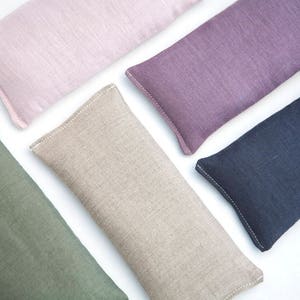 May include: Five rectangular linen eye pillows in different colors: pink, purple, beige, green, and dark blue. The pillows are all sewn with white thread and have a subtle texture.