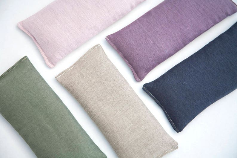 May include: Five rectangular linen eye pillows in different colors: pink, purple, beige, green, and dark blue. The pillows are all sewn with white thread and have a subtle texture.