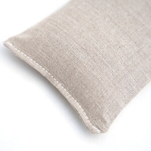 May include: A beige linen eye pillow with white stitching. The pillow is rectangular and has a slightly rounded shape.