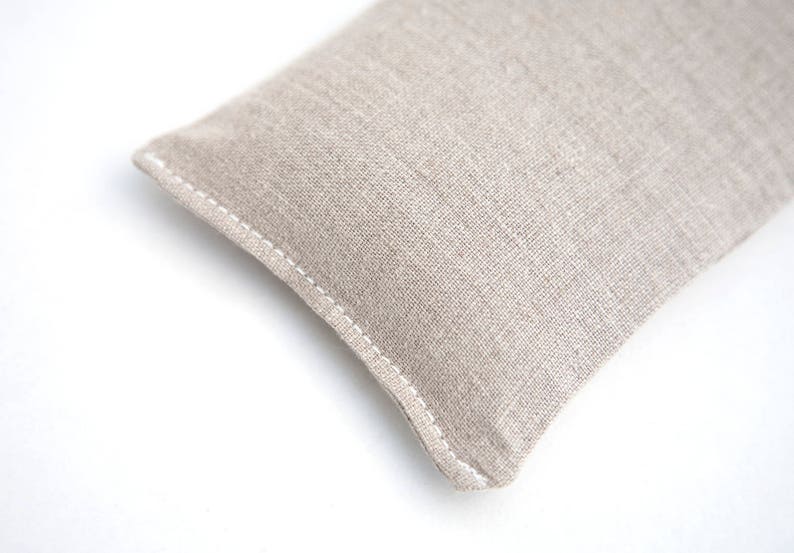 May include: A beige linen eye pillow with white stitching. The pillow is rectangular and has a slightly rounded shape.