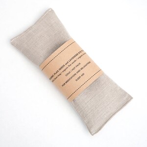 May include: A beige linen eye pillow filled with flax seeds and lavender. The pillow has a brown paper label that reads "Organic Flax Seeds and Lavender Eye Pillow", "100% linen bag / organic flax seeds / organic lavender", "Cold / Hot Pack", "For Meditation and Relaxation", and "Sleep Aid".