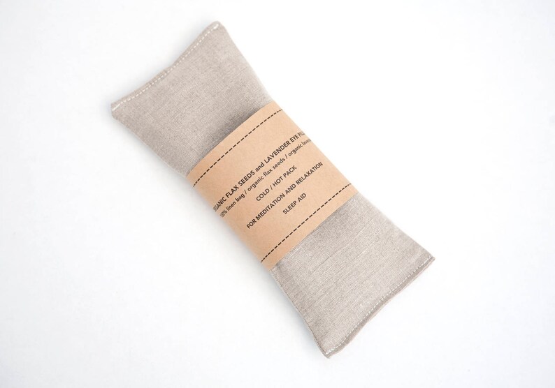 May include: A beige linen eye pillow filled with flax seeds and lavender. The pillow has a brown paper label that reads "Organic Flax Seeds and Lavender Eye Pillow", "100% linen bag / organic flax seeds / organic lavender", "Cold / Hot Pack", "For Meditation and Relaxation", and "Sleep Aid".