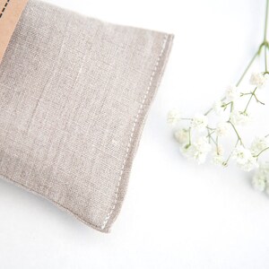 May include: A light brown linen eye pillow with the text "Sleep Aid" on the brown paper label. The pillow is on a white surface with a sprig of white baby's breath flowers.