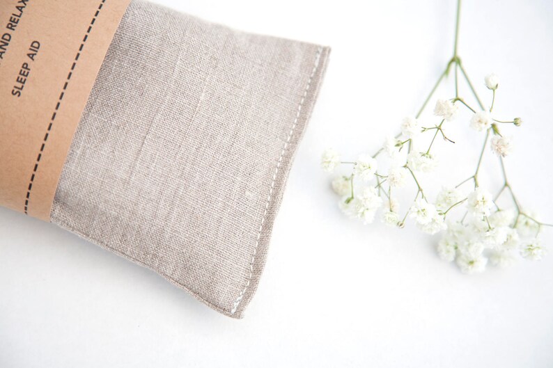 May include: A light brown linen eye pillow with the text "Sleep Aid" on the brown paper label. The pillow is on a white surface with a sprig of white baby's breath flowers.