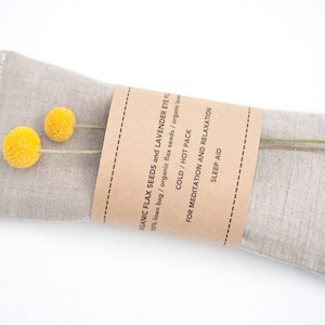 May include: A beige linen eye pillow filled with organic flax seeds and lavender. The pillow has a brown paper label that reads "ORGANIC FLAX SEEDS and LAVENDER EYE PILLOW 100% linen bag / organic flax seeds / organic COLD / HOT PACK FOR MEDITATION AND RELAXATION SLEEP AID".