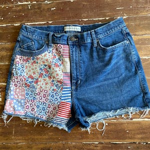 May include: Blue denim shorts featuring a high-waisted design with a button and zipper closure. The left side panel is embellished with a patchwork of floral, polka dot, and striped fabric patterns. The hemline has a frayed, raw edge finish.