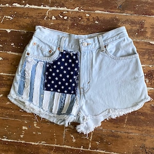 May include: Light blue denim shorts with a distressed hem. The left side features a patchwork design of the American flag, with blue and white stripes and a navy blue field of white stars. The shorts have a classic five-pocket design.