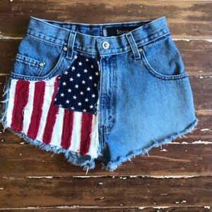 May include: Blue denim shorts with a frayed hem and a red, white, and blue American flag patch on the front. The shorts have a loose fit and a high waist.