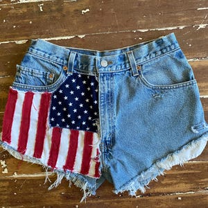 May include: Light blue denim shorts featuring a high-waisted cut with a metal button closure and belt loops. The front left panel is embellished with a fabric patch displaying the American flag, while the leg openings have a frayed hem finish.