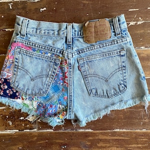 May include: Light blue denim shorts featuring a frayed hem and a brown leather patch on the back waistband. The left side is accented with a multi-colored patchwork fabric panel displaying floral, paisley, and plaid patterns in blue, pink, and white.