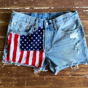May include: Light blue denim shorts with a distressed look. The shorts feature a patch resembling the American flag on the front, with red and white stripes and a blue field of white stars. The shorts have a button closure and pockets.