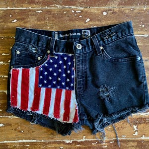 May include: Black denim shorts featuring a distressed finish and frayed hem. The front left panel is accented with a sewn-on patch displaying the American flag in red, white, and blue. The interior waistband label reads hipster boot co.