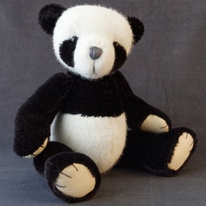 May include: A black and white plush panda bear toy with a black nose and black eyes. The panda is sitting with its legs crossed and its paws resting on its lap.